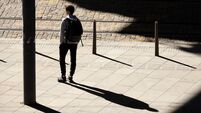One teenage boy standing alone waiting for a tram on bus stop in sunlight, with shadows