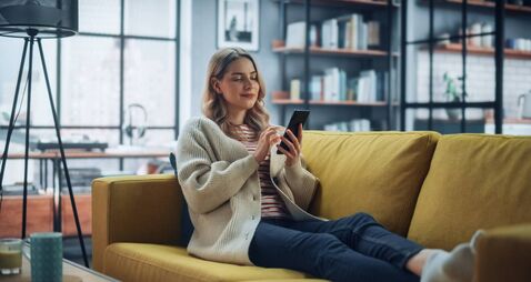 Beautiful Caucasian Female Using Smartphone in Stylish Living Room while Resting on a Cozy Couch Sofa. Young Woman at Home, Brow
