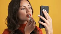Close up beautiful brunette girl applying red lipstick looking in smartphone over colorful background